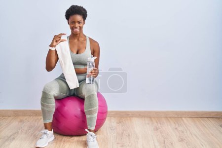Foto de African american woman wearing sportswear sitting on pilates ball smiling and confident gesturing with hand doing small size sign with fingers looking and the camera. measure concept. - Imagen libre de derechos