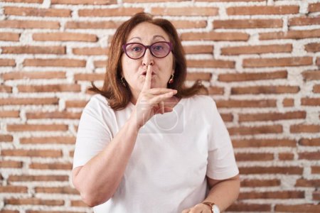 Téléchargez les photos : Femme âgée avec des lunettes debout sur un mur de briques demandant de se taire avec le doigt sur les lèvres. silence et concept secret. - en image libre de droit