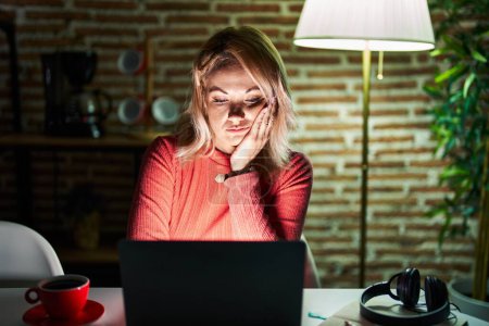 Téléchargez les photos : Femme blonde utilisant un ordinateur portable la nuit à la maison pensant fatigué et ennuyé avec des problèmes de dépression avec les bras croisés. - en image libre de droit