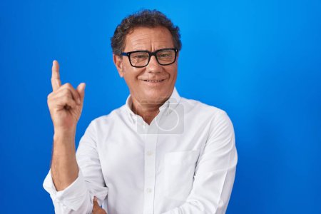 Foto de Middle age hispanic man standing over blue background with a big smile on face, pointing with hand finger to the side looking at the camera. - Imagen libre de derechos