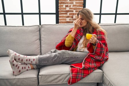 Foto de Young woman sitting on the sofa drinking a coffee at home tired rubbing nose and eyes feeling fatigue and headache. stress and frustration concept. - Imagen libre de derechos