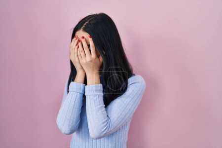 Foto de Mujer hispana de pie sobre fondo rosa con expresión triste cubriendo la cara con las manos mientras llora. concepto de depresión. - Imagen libre de derechos
