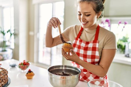 Téléchargez les photos : Jeune belle femme hispanique souriant confiant mettre du chocolat sur muffin à la cuisine - en image libre de droit