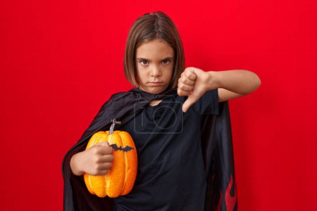 Foto de Pequeño niño hispano usando una capa y sosteniendo la calabaza de Halloween con la cara enojada, signo negativo que muestra aversión con los pulgares hacia abajo, concepto de rechazo - Imagen libre de derechos