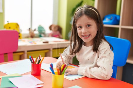 Téléchargez les photos : Adorable hispanic girl preschool student sitting on table drawing on paper at kindergarten - en image libre de droit