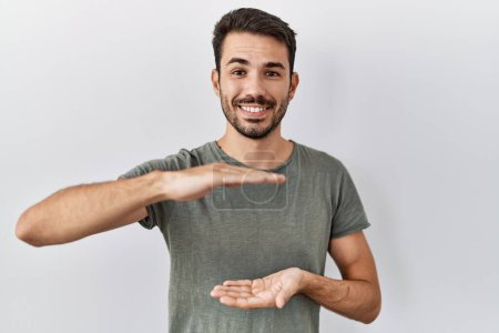 Foto de Young hispanic man with beard wearing casual t shirt over white background gesturing with hands showing big and large size sign, measure symbol. smiling looking at the camera. measuring concept. - Imagen libre de derechos