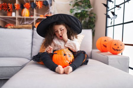 Foto de Adorable hispanic girl wearing halloween costume sitting on sofa with surprise expression at home - Imagen libre de derechos