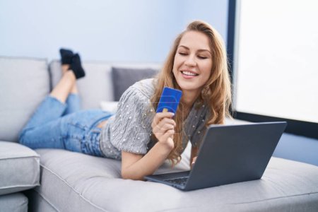 Foto de Mujer rubia joven usando el ordenador portátil y la tarjeta de crédito tumbado en el sofá en casa - Imagen libre de derechos