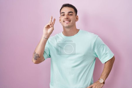 Téléchargez les photos : Bel homme hispanique debout sur fond rose souriant regardant la caméra montrant doigts faisant signe de victoire. numéro deux. - en image libre de droit