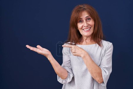 Téléchargez les photos : Middle age woman standing over blue background amazed and smiling to the camera while presenting with hand and pointing with finger. - en image libre de droit