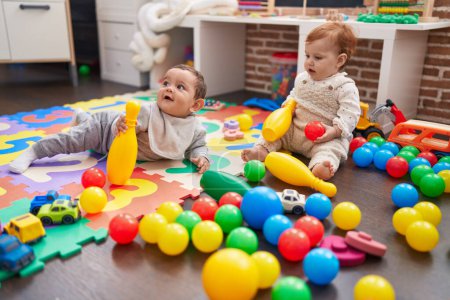 Foto de Dos adorables bebés jugando con pelotas y bolos sentados en el suelo en el jardín de infantes - Imagen libre de derechos