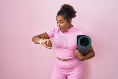 Téléchargez les photos : Young hispanic woman with curly hair holding yoga mat over pink background looking at the watch time worried, afraid of getting late - en image libre de droit