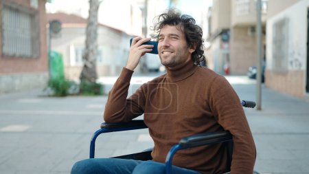 Photo for Young hispanic man listening voice message by smartphone sitting on wheelchair at street - Royalty Free Image