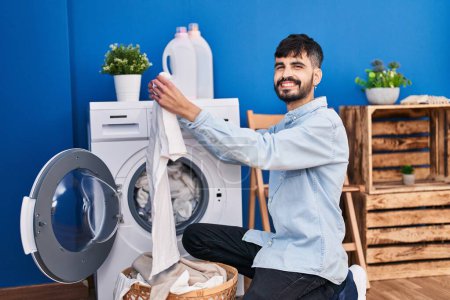 Téléchargez les photos : Jeune homme hispanique tenant t-shirt blanc laver les vêtements à la buanderie - en image libre de droit