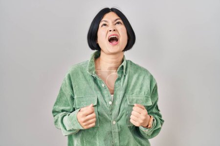 Foto de Mujer asiática joven de pie sobre fondo blanco enojado y loco gritando frustrado y furioso, gritando con ira. rabia y concepto agresivo. - Imagen libre de derechos