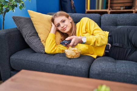 Foto de Young blonde woman watching tv lying on sofa at home - Imagen libre de derechos
