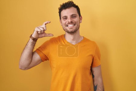 Téléchargez les photos : Jeune homme hispanique debout sur fond jaune geste souriant et confiant avec la main faisant signe de petite taille avec les doigts regardant et la caméra. concept de mesure. - en image libre de droit