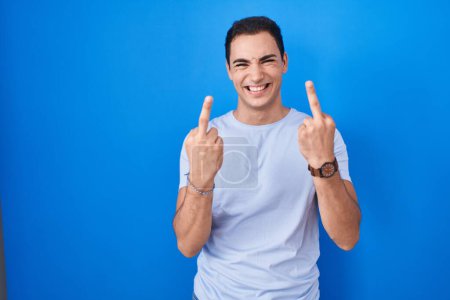 Foto de Young hispanic man standing over blue background showing middle finger doing fuck you bad expression, provocation and rude attitude. screaming excited - Imagen libre de derechos