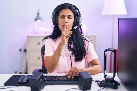 Photo for Mature hispanic woman playing video games at home asking to be quiet with finger on lips. silence and secret concept. - Royalty Free Image