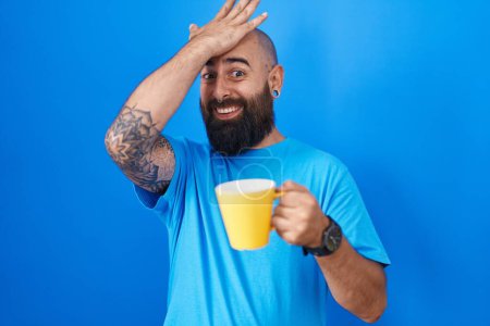 Téléchargez les photos : Jeune homme hispanique avec barbe et tatouages boire une tasse de café surpris avec la main sur la tête pour erreur, rappelez-vous erreur. oublié, mauvais concept de mémoire. - en image libre de droit