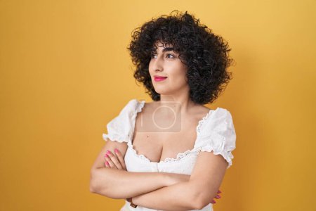 Téléchargez les photos : Jeune femme brune aux cheveux bouclés debout sur fond jaune regardant sur le côté avec les bras croisés convaincus et confiants - en image libre de droit