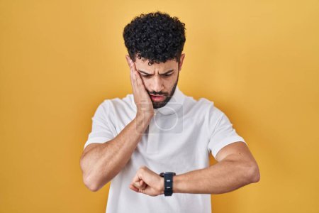 Téléchargez les photos : Homme arabe debout sur fond jaune regardant l'heure de la montre inquiet, peur de se faire tard - en image libre de droit