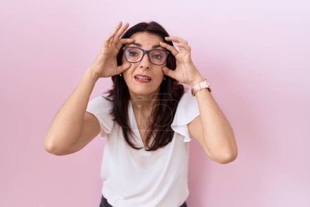Téléchargez les photos : Femme hispanique du Moyen Âge portant un t-shirt blanc décontracté et des lunettes essayant d'ouvrir les yeux avec les doigts, endormie et fatiguée pour la fatigue matinale - en image libre de droit