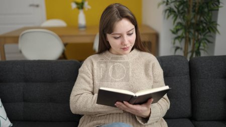 Téléchargez les photos : Young blonde woman reading book sitting on sofa at home - en image libre de droit