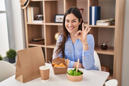 Téléchargez les photos : Young brunette woman eating take away food at home doing ok sign with fingers, smiling friendly gesturing excellent symbol - en image libre de droit
