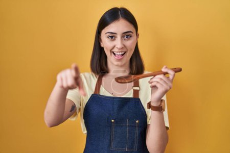 Foto de Chica hispana comiendo cuchara de madera saludable señalándote a ti y a la cámara con los dedos, sonriendo positiva y alegre - Imagen libre de derechos