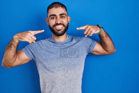 Téléchargez les photos : Moyen-Orient homme avec barbe debout sur fond bleu souriant gai montrant et pointant avec les doigts dents et la bouche. concept de santé dentaire. - en image libre de droit