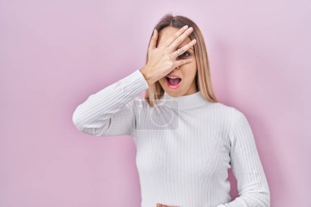 Foto de Mujer rubia joven con suéter blanco sobre fondo rosa asomándose en shock cubriendo la cara y los ojos con la mano, mirando a través de los dedos con expresión avergonzada. - Imagen libre de derechos
