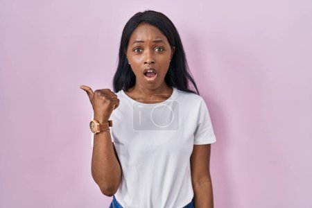 Photo for African young woman wearing casual white t shirt surprised pointing with hand finger to the side, open mouth amazed expression. - Royalty Free Image