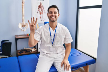 Photo for Young hispanic man with beard working at pain recovery clinic showing and pointing up with fingers number four while smiling confident and happy. - Royalty Free Image