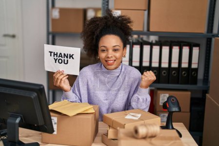 Téléchargez les photos : Young african american woman working at small business ecommerce holding thank you banner screaming proud, celebrating victory and success very excited with raised arm - en image libre de droit