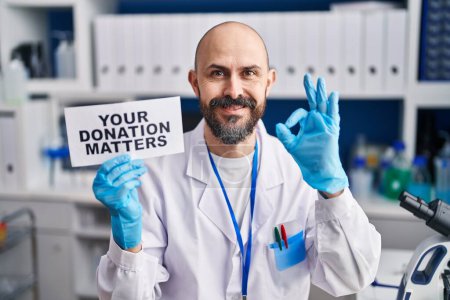 Téléchargez les photos : Jeune homme hispanique travaillant au laboratoire scientifique vous tenant don questions bannière faire ok signe avec les doigts, sourire geste amical excellent symbole - en image libre de droit