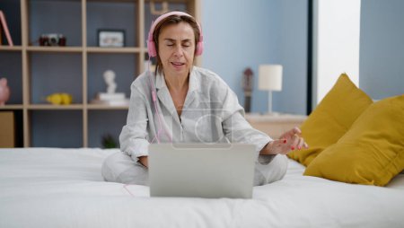 Téléchargez les photos : Moyen Age femme hispanique écouter de la musique assis sur le lit dans la chambre - en image libre de droit