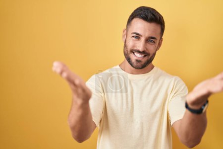 Foto de Hombre hispano guapo de pie sobre fondo amarillo sonriente alegre ofreciendo manos dando asistencia y aceptación. - Imagen libre de derechos