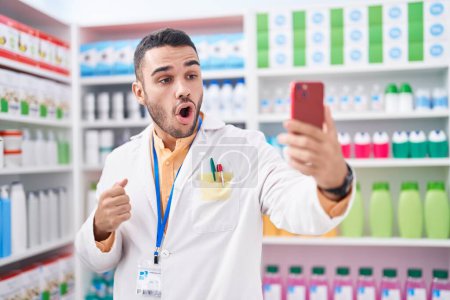 Téléchargez les photos : Young hispanic man working at pharmacy drugstore doing video call with smartphone scared and amazed with open mouth for surprise, disbelief face - en image libre de droit
