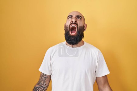 Photo for Young hispanic man with beard and tattoos standing over yellow background angry and mad screaming frustrated and furious, shouting with anger. rage and aggressive concept. - Royalty Free Image