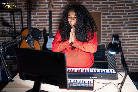 Photo for Plus size hispanic woman playing piano at music studio praying with hands together asking for forgiveness smiling confident. - Royalty Free Image