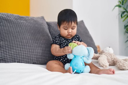 Téléchargez les photos : Adorable bébé hispanique jouant avec des poupées assises sur le lit dans la chambre - en image libre de droit