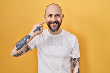 Foto de Joven hombre hispano con tatuajes de pie sobre fondo amarillo señalando con la mano el dedo a la cara y la nariz, sonriendo alegre. concepto de belleza - Imagen libre de derechos
