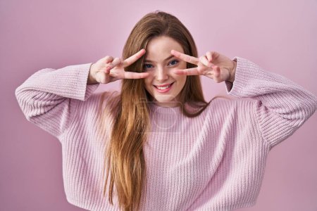 Foto de Joven mujer caucásica de pie sobre fondo rosa haciendo símbolo de paz con los dedos sobre la cara, sonriendo alegre mostrando la victoria - Imagen libre de derechos