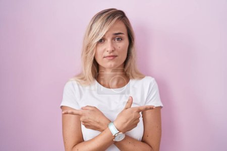 Téléchargez les photos : Jeune femme blonde debout sur fond rose pointant des deux côtés avec les doigts, direction différente en désaccord - en image libre de droit