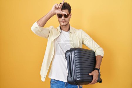 Téléchargez les photos : Jeune homme hispanique tenant une valise allant en vacances d'été se moquant des gens avec les doigts sur le front faisant geste de perdant se moquant et insultant. - en image libre de droit