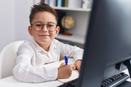 Photo for Adorable hispanic boy student using computer writing on notebook at classroom - Royalty Free Image