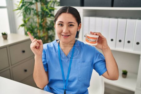 Téléchargez les photos : Jeune dentiste latino-américaine tenant une prothèse dentaire avec des bretelles souriantes heureux pointant avec la main et le doigt sur le côté - en image libre de droit