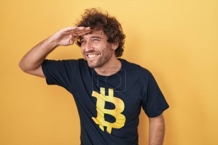 Photo for Hispanic young man wearing bitcoin t shirt very happy and smiling looking far away with hand over head. searching concept. - Royalty Free Image