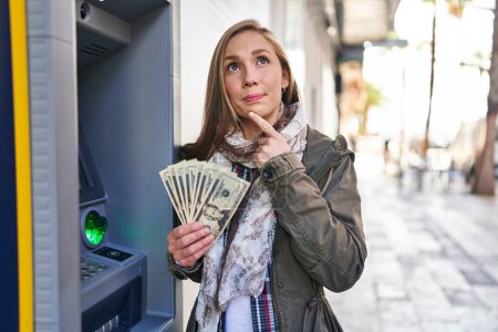 Foto de Mujer rubia joven sosteniendo billetes de dólares de cajero automático cara seria pensando en la pregunta con la mano en la barbilla, reflexivo acerca de la idea confusa - Imagen libre de derechos
